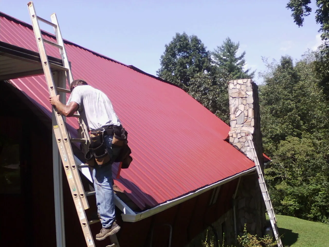 Roofer on a ladder beside a red metal roof and stone chimney — Soffit & Fascia Repair by Roofing Brothers in Freeburg